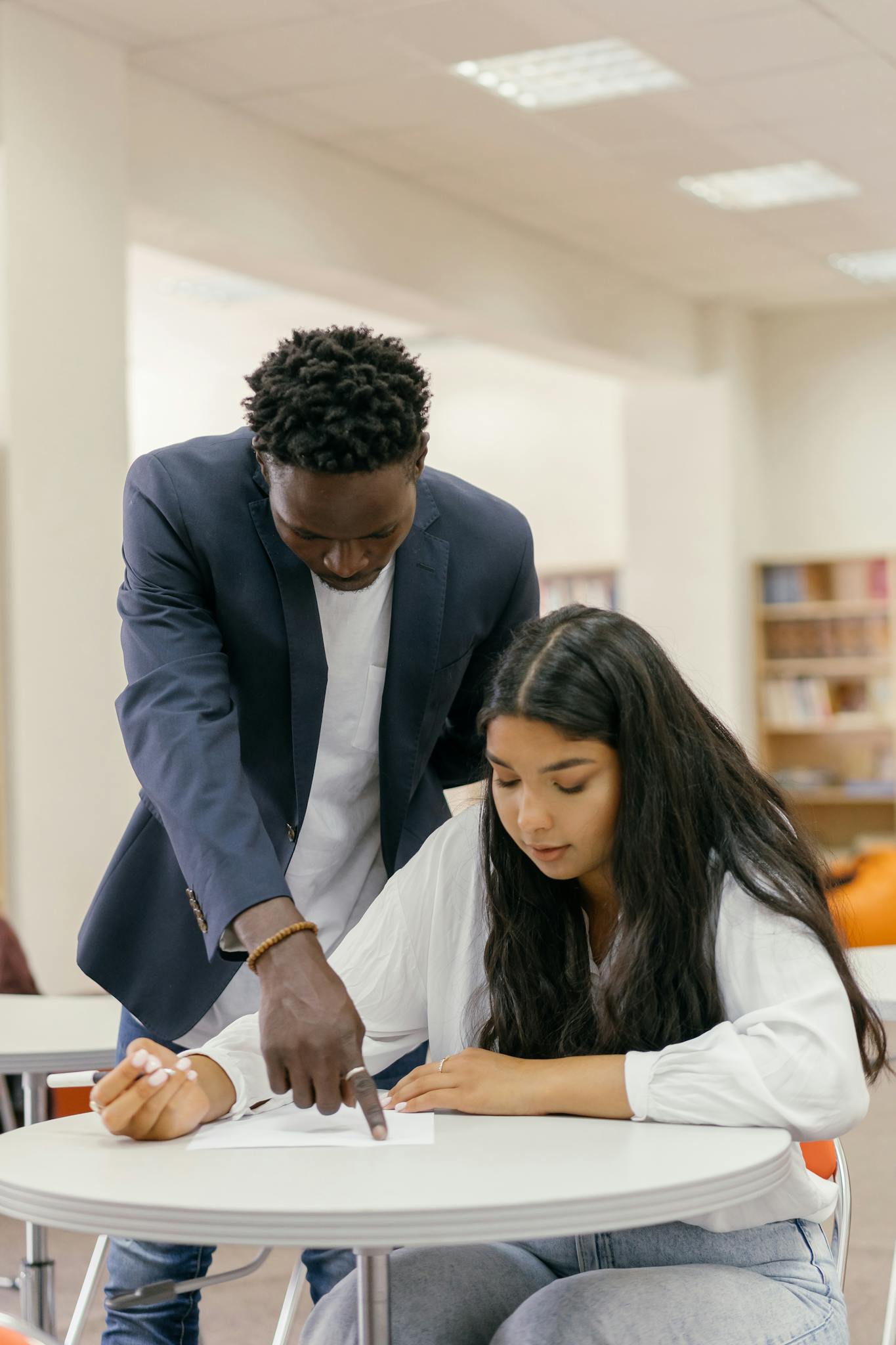 A man tutor guides a woman student during a study session in a library setting.