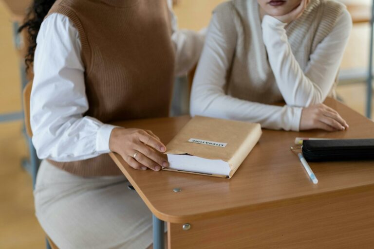 Two students sitting together at a classroom desk with a book titled 'Literature' and stationery items.